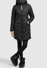 Abrigo acolchado negro con capucha, con costuras diagonales, cremallera frontal y bolsillos laterales. Combinado con leggings negros y botas.