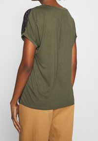 Olive green t-shirt with rolled sleeves, featuring a textured black patterned panel on the shoulders and soft fabric.