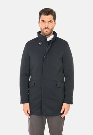 Uomo che indossa un cappotto blu scuro con collo alto, tasche frontali e bottoni a gommino, in piedi contro uno sfondo chiaro e semplice.