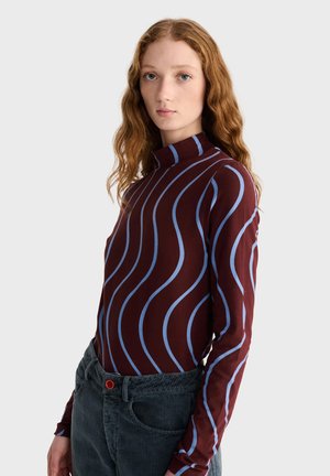 Jeune femme aux longs cheveux rouges ondulés portant un col roulé bordeaux à rayures ondulées bleu clair et un pantalon en velours côtelé foncé avec un bouton rouge.