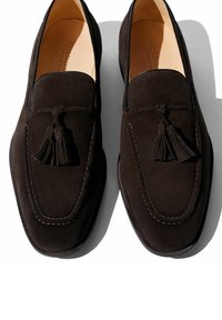 Bruine suede instappers hebben een moccasin-stijl ontwerp met stikdetails en dubbele kwasten aan de voorkant. De gladde leren voering maakt het geheel af.