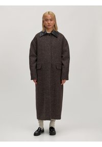 Langer brauner Mantel aus strukturiertem Stoff mit schwarzem Lederkragen. Verfügt über seitliche Taschen und einen geraden Schnitt. Das Modell trägt schwarze Schuhe und beige Socken.
