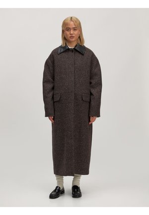 Lang bruin jasje van gestructureerde stof met een zwarte leren kraag. Heeft zijzakken en een rechte snit. Het model draagt zwarte schoenen en beige sokken.