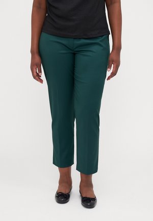 Nainen, joka on pukeutunut tummanvihreisiin nilkkapituisiiin housuihin ja mustiin balettitossuihin, seisoo yksinkertaisen valkoisen taustan edessä.