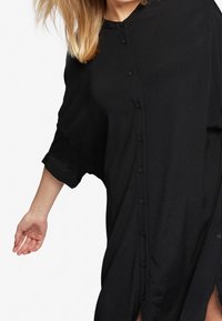 Svart knäppklänning med korta ärmar och sidokilar, tillverkad av lättviktig tyg, med en avslappnad passform och subtil struktur.