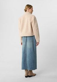 Giacca in pile rosa chiaro con una texture morbida, design corto e colletto, abbinata a una lunga gonna di jeans blu e stivaletti beige alla caviglia.