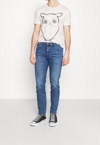 Herrens outfit med en ljus beige t-shirt med en ugglegrafik, i kombination med åtsittande blå jeans och svarta sneakers med vita sulor.