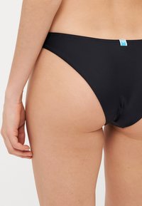 Black bikini bottoms made of smooth, stretchy fabric. Features a low-rise cut and a small blue tag on the back right side.