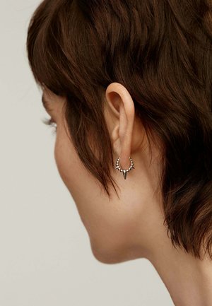 Pendiente de aro plateado con acentos puntiagudos y pequeños detalles de cuentas, llevado en una oreja de cabello castaño oscuro y corto.