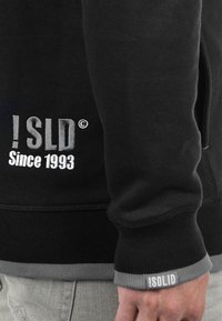 Fekete pulóver hímzett "!SLD" logóval és "Since 1993" felirattal szürkében. Texturált anyag, bordázott mandzsettákkal és aljjal, szürke díszítéssel.