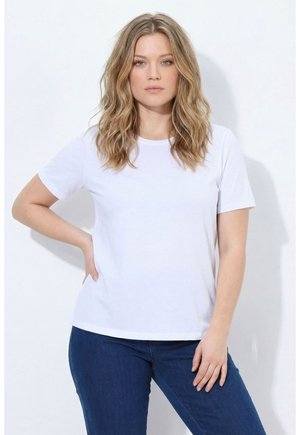 Frau mit welligem blondem Haar trägt ein schlichtes weißes Kurzarm-T-Shirt und dunkelblaue Jeans, posiert mit einer Hand in der Hüfte vor weißem Hintergrund.