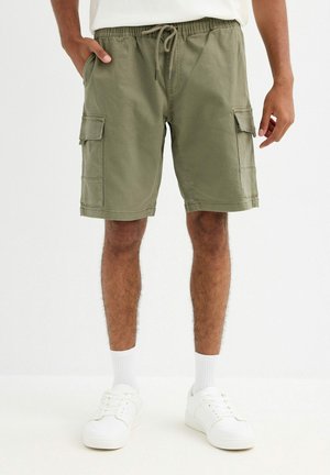 Hombre vistiendo pantalones cortos tipo cargo verde oliva con cintura de cordón, calcetines blancos y zapatillas blancas, de pie frente a un fondo liso.