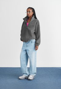 Giacca in pile grigia con zip e colletto alto, abbinata a jeans azzurri a gamba larga e scarpe da ginnastica grigie, con una maglietta rosa sotto.