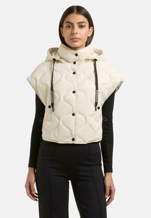 Frau trägt eine ärmellose, gesteppte cremefarbene Jacke mit Kapuze und schwarzen Kordeln über einem schwarzen Langarmshirt und schwarze Hose.