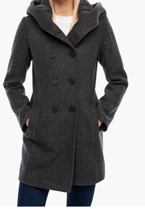 Manteau en laine gris foncé à double boutonnage avec large col et mains dans les poches, porté par-dessus un haut blanc et un jean bleu.