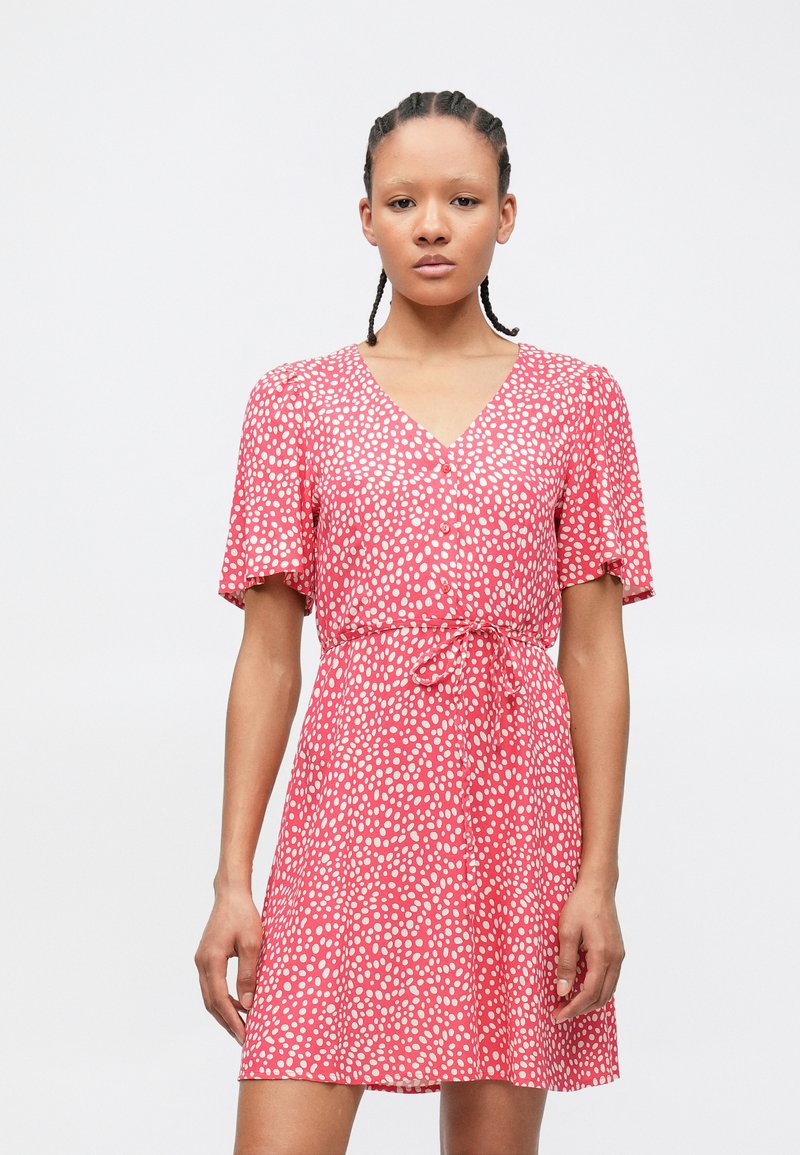 Γυναίκα που φοράει ροζ φόρεμα με κοντά μανίκια, λευκές πουά, κουμπιά στο μπροστινό μέρος και ζωνάκι στη μέση, στέκεται μπροστά σε ένα απλό φόντο.