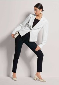 Weiß strukturierter Blazer mit langen Ärmeln, schwarzer enganliegenden Bluse, schwarze Hose und hellbeige Blockabsatzschuhe mit einer Schleifenverzierung.