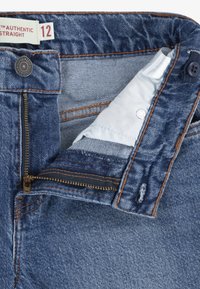 Jean en denim bleu avec fermeture éclair et bouton partiellement ouverts, montrant la doublure intérieure blanche et une étiquette de taille indiquant 12 avec des détails de couture orange.