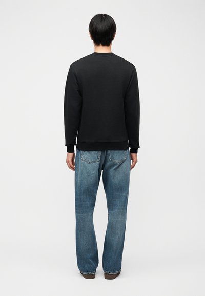 Άτομο που στέκεται γυρισμένο με την πλάτη, φορώντας μαύρο πουκάμισο με μακριά μανίκια, μπλε τζιν και καφέ παπούτσια, μπροστά σε απλό φωτεινό φόντο.