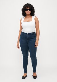 White square-neck top with fitted design, paired with high-waisted dark blue skinny jeans. Black sunglasses and flat shoes complete the look.