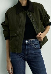 Veste courte vert foncé avec un col en velours côtelé, fermeture éclair, et poches boutonnées, associée à un jean bleu foncé et un haut noir.