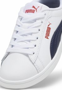 Białe sneakersy z granatowymi akcentami, okrągły przód, gładki syntetyczny materiał i płaskie białe sznurówki. Zawierają logo PUMA w kolorze czerwonym.