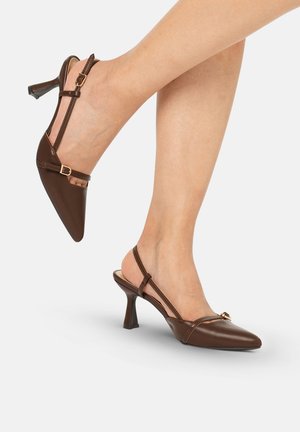 Tacones slingback de cuero marrón con punta afilada y pequeñas hebillas doradas, usados con las piernas cruzadas sobre un fondo blanco.