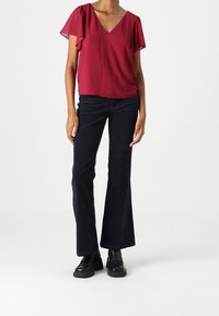 Bordeaux blouse met zwierige mouwen en v-hals; gecombineerd met flared zwarte broeken en chunky zwarte schoenen. Glad materiaal en aansluitend ontwerp.