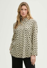 Hemd in Creme mit schwarzem Zickzackmuster, langen Ärmeln, Kragen und seitlichen Taschen, aus weichem Stoff gefertigt.