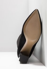 Chaussure à talon haut en daim noir avec bout pointu, semelle noire texturée et talon bloc géométrique. Semelle intérieure claire avec des rainures.