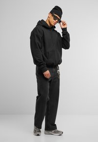 Sweatshirt à capuche noire avec poche kangourou, jean denim noir, accessoires en métal argenté et chaussures de sport grises. Les accessoires incluent des lunettes de soleil noires et un bonnet.