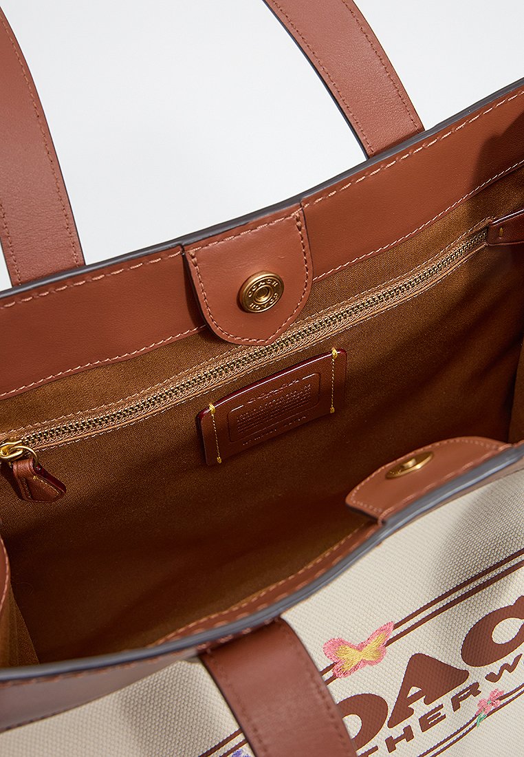 Bolsa de lona beige con acentos de cuero marrón, que cuenta con un cierre magnético, un bolsillo interior con cremallera y detalles del logo.