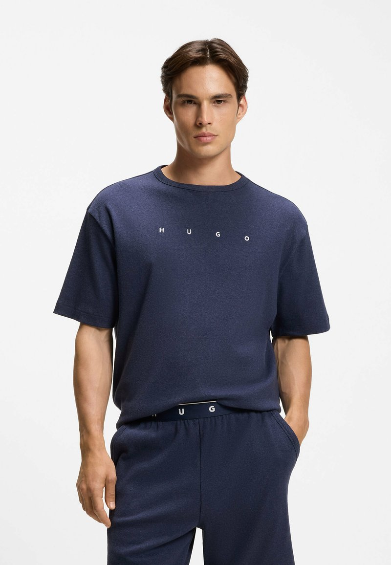 Jeune homme debout, mains dans les poches, portant un t-shirt bleu marine HUGO et un pantalon assorti sur fond blanc.