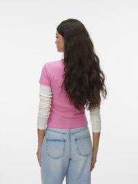 Γυναίκα με μακριά, σκούρα, κυματιστά μαλλιά φοράει ροζ μπλούζα με κοντά μανίκια και στρώσεις πάνω από λευκά μακριά μανίκια και ανοιχτόχρωμα μπλε τζιν, στέκεται με την πλάτη στραμμένη προς τα εμπρός.