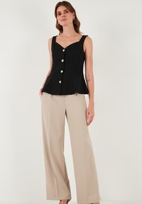 Schwarzes Peplum-Top mit dünnen Trägern, das vorne mit sechs metallischen Knöpfen verziert ist. Kombiniert mit beigen High-Waist-Hosen. Glatte Stoffstruktur.