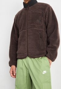 Brun fleecejacka med hög krage, dragkedja framtill och bröstficka; har textilerad tyg och kontrastdragkedjor.