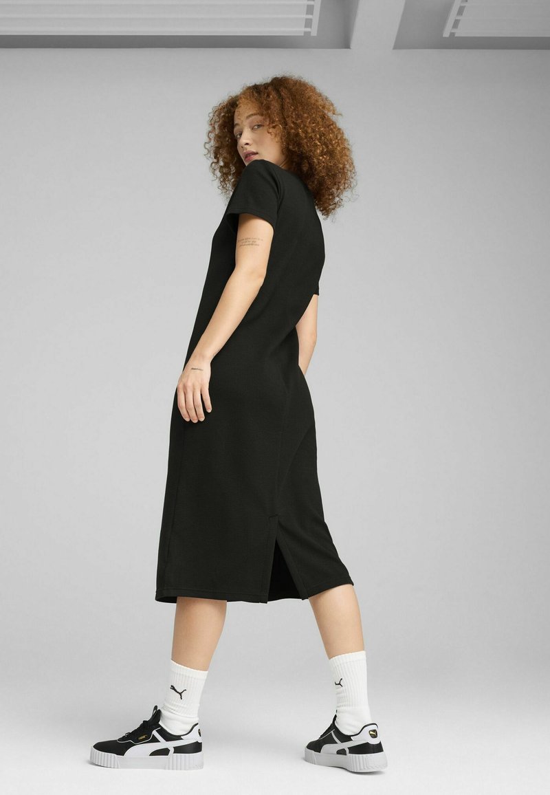 Robe noire mi-longue à manches courtes, avec une fente sur le côté. Associée à des chaussures de sport noires et des chaussettes blanches.