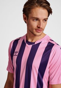 Lila och mörkpurpur randig sporttröja med korta ärmar, V-halsdesign och en logotyp på vänster sida. Tillverkad av ett mjukt tyg.