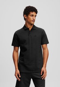 Cămașă polo neagră dintr-un material respirabil, cu guler clasic, mâneci scurte, închidere cu trei nasturi și broderie discretă cu logo.