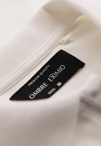 Vit tyg med en synlig svart etikett. Etiketten har "OMBRE EXIMIO" i silverfärgade bokstäver och anger storlek M. Texturerad sömnad längs kanterna.