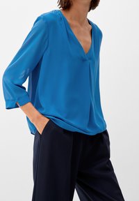 Femme portant un chemisier bleu ample à col en V avec manches trois-quarts et un pantalon large foncé, les mains dans les poches, sur un fond uni.