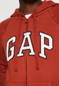 Close-up van een persoon die een roestrode zip-up hoodie draagt met grote witte letters met donkere randen die het woord "GAP" over de borst spellen.