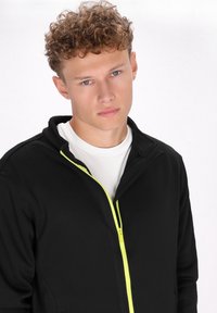Giacca nera con zip, dotata di un colletto alto e un zip giallo brillante. Il tessuto appare liscio e leggero, indossata sopra una camicia bianca.
