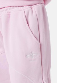 Pantalones de chándal rosa claro con bolsillos laterales, una textura suave y un logo bordado en blanco en el muslo con detalles de costura curva.