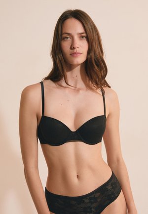 Mujer con cabello largo y castaño que lleva un sujetador de encaje negro y ropa interior de encaje a juego sobre un fondo beige.