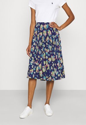 Kvinde iført en mørkeblå blomsterprintet plisseret midi-nederdel, hvid T-shirt med korte ærmer og hvide sneakers, står med den ene hånd på hoften.