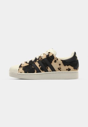 Sneaker mit Rindsledermuster in Beige und Schwarz, drei schwarze Streifen, weiße Gummikappe und strukturierte Sohle. Verfügt über Schnürsenkel und Branding.