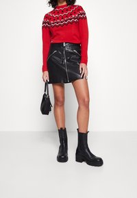 Rode trui met een patroon aan de schouders, zwarte leren mini-jurk met ritsdetails, en zwarte enkelboots met dikke zolen. Zwart handtasje inbegrepen.