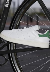Białe skórzane sneakersy z zielonymi akcentami, z perforowanymi detalami i teksturowaną podeszwą, umieszczone na tle czarnej ramy roweru.