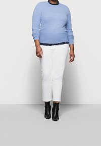 Hellblauer, strukturierter Pullover über einem marineblauen, gemusterten Hemd, kombiniert mit weißen, knöchellangen Hosen und schwarzen Stiefeletten.